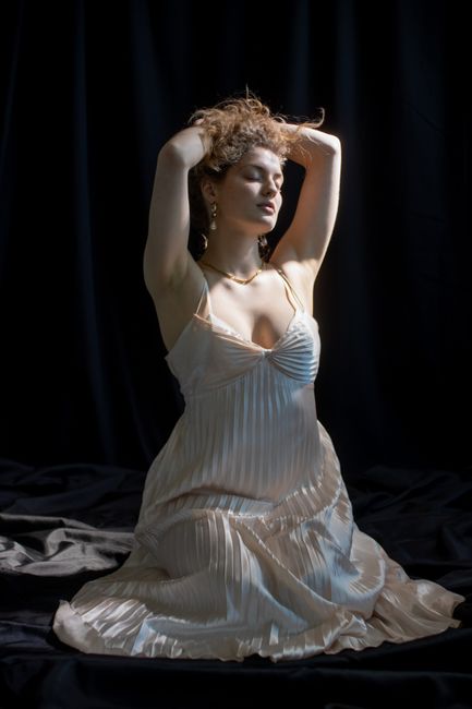 Charming female with long hair sunlit face and closed eyes wearing long beige dress with bare shoulders and chest touching head while sitting against dark blue background