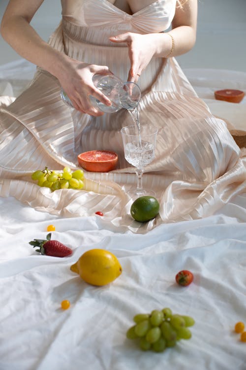 High angle of crop anonymous female in elegant dress pouring water from bottle into glass while sitting on white blanket near scattered assorted fresh fruits