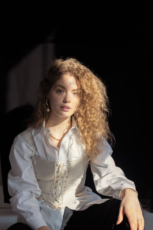 Free Confident young female model with curly blond hair in elegant white shirt and corset sitting in studio and looking at camera against black background Stock Photo