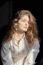 Dreamy young female model with curly blond hair in stylish corset and white shirt looking away pensively against black background in daylight