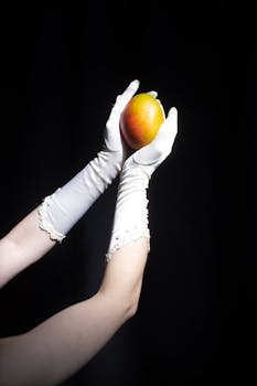 Crop unrecognizable lady in long elegant white gloves demonstrating fresh ripe mango against black background