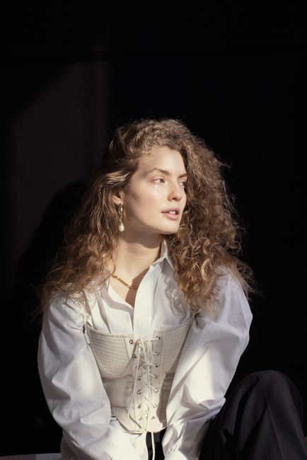 Confident young female millennial with curly blond hair looking away in stylish white corset and shirt looking away dreamily in dark room