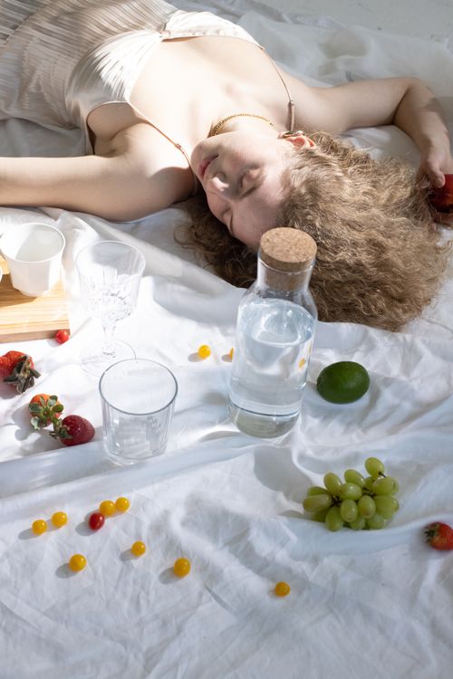 Free From above of feminine young lady with blond hair in stylish dress lying on white bed sheet with closed eyes near water glass bottle and scattered assorted fruits in sunlight Stock Photo
