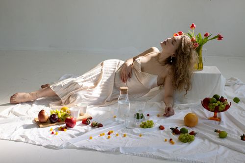 Free Full body of young tranquil barefooted lady in elegant silk dress lying on blanket on floor near vase with fresh tulips and assorted ripe colorful fruits Stock Photo
