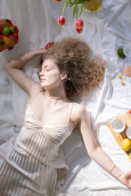 Top view of graceful young female with curly blond hair in stylish light dress lying on white textile with closed eyes near fresh tulips and fruits in sunlight