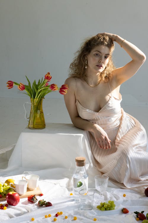 Elegant young woman in maxi dress touching curly blond hair and looking at camera while sitting in floor and leaning on small table with vase of tulips amidst scattered fruits