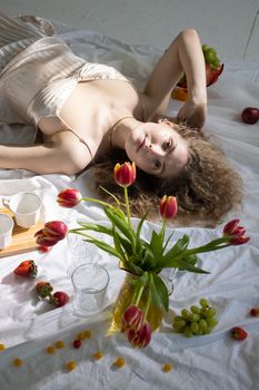 High angle of sensual woman with curly hair in silk dress relaxing on floor near to vase with flowers and coffee cups and scattered fruits