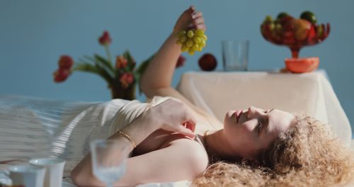 Free Side view of calm female with curly hair in dress lying with grape in hand and looking away against table with flowers and fruits Stock Photo