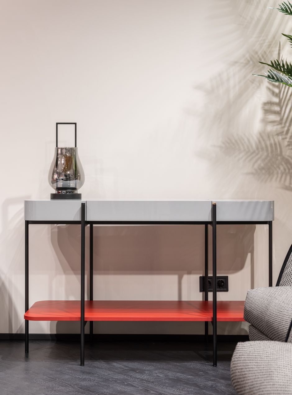 Modern table with decorative vase and red shelf placed at white wall in light room with gray seat and green plant