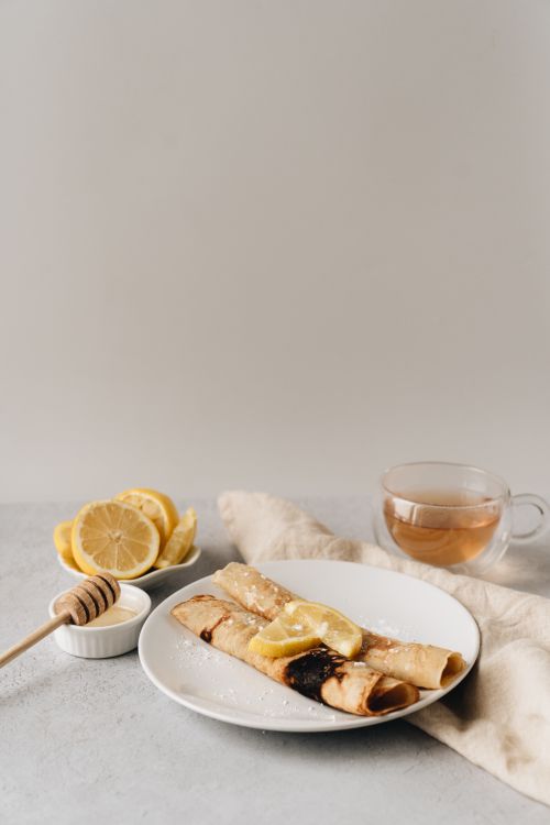 Free A delightful breakfast setup featuring crêpes with honey, lemon slices, and a cup of tea, perfect for a cozy morning. Stock Photo