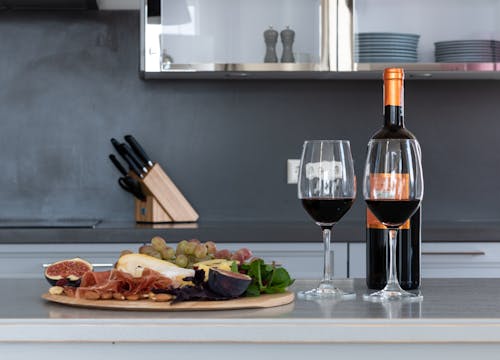 Goblets with red wine placed on counter with bottle of alcohol drink and tray with assorted fruits and meat in kitchen