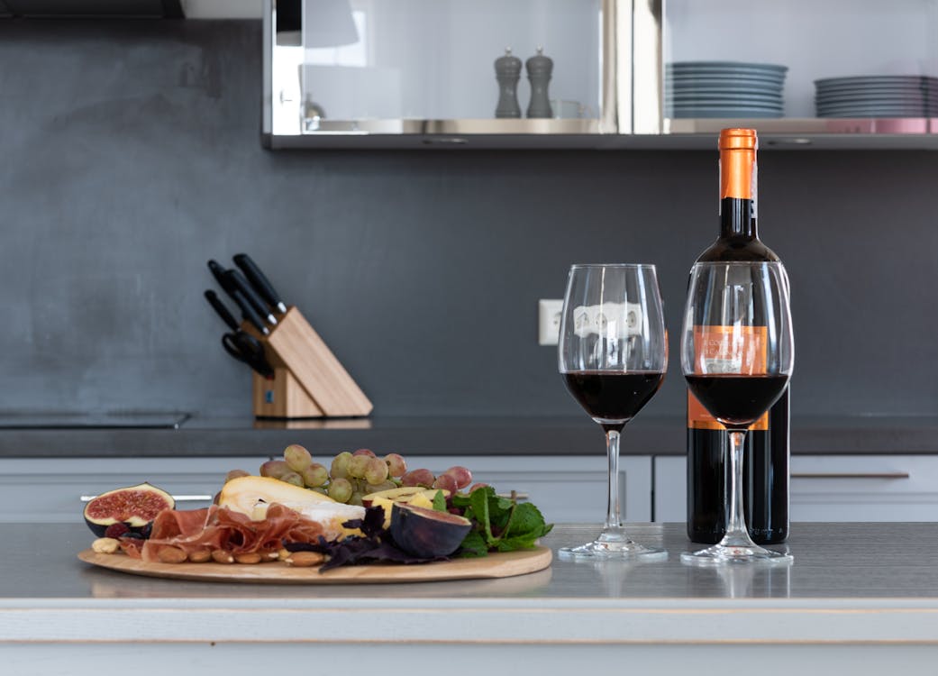 Free Goblets with red wine placed on counter with bottle of alcohol drink and tray with assorted fruits and meat in kitchen Stock Photo