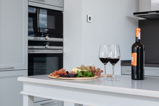 Goblets of red wine placed on white counter with bottle of alcohol beverage and tray of assorted appetizers in kitchen with modern appliances