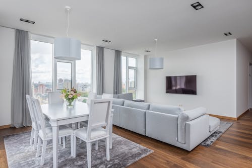 Vase with flowers on white table near chairs placed on rug near couch against wall with TV in stylish apartment with big windows