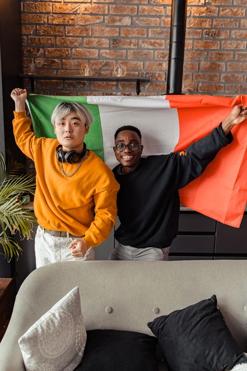 Free Two friends celebrate together holding an Italian flag inside a cozy room. Stock Photo