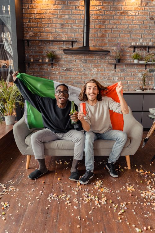 Free Two smiling friends sitting on a sofa holding drinks and an Italian flag, celebrating indoors. Stock Photo