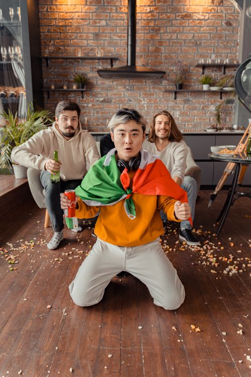 Free Three friends indoors, one kneeling with a flag, celebrating and enjoying drinks together. Stock Photo