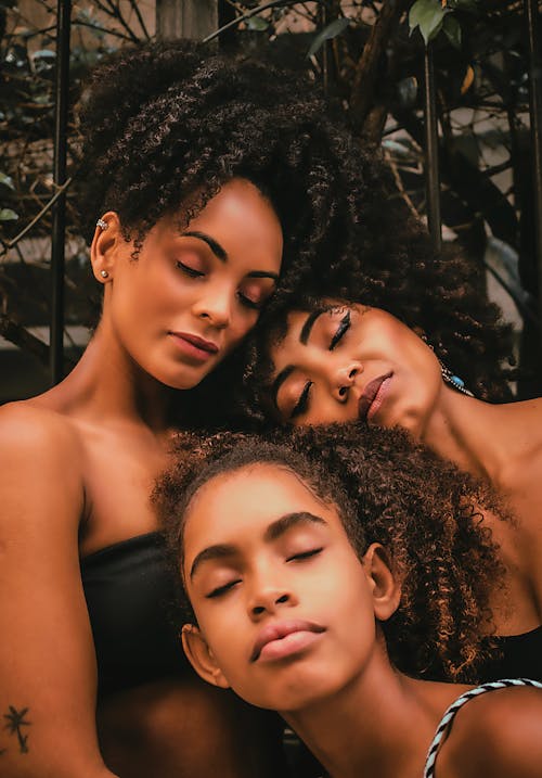 Multi generational African American ladies and girl with closed eyes and curly hair leaning head on each other in daytime in street
