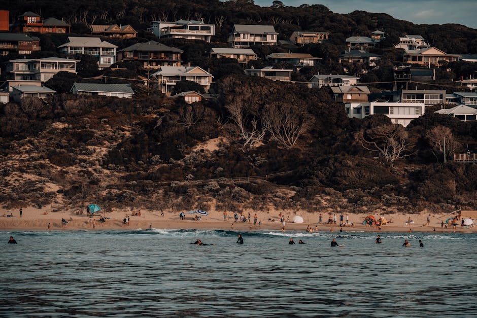 Idyllic beach scene with surfers and hillside vacation homes overlooking a sandy shore.
