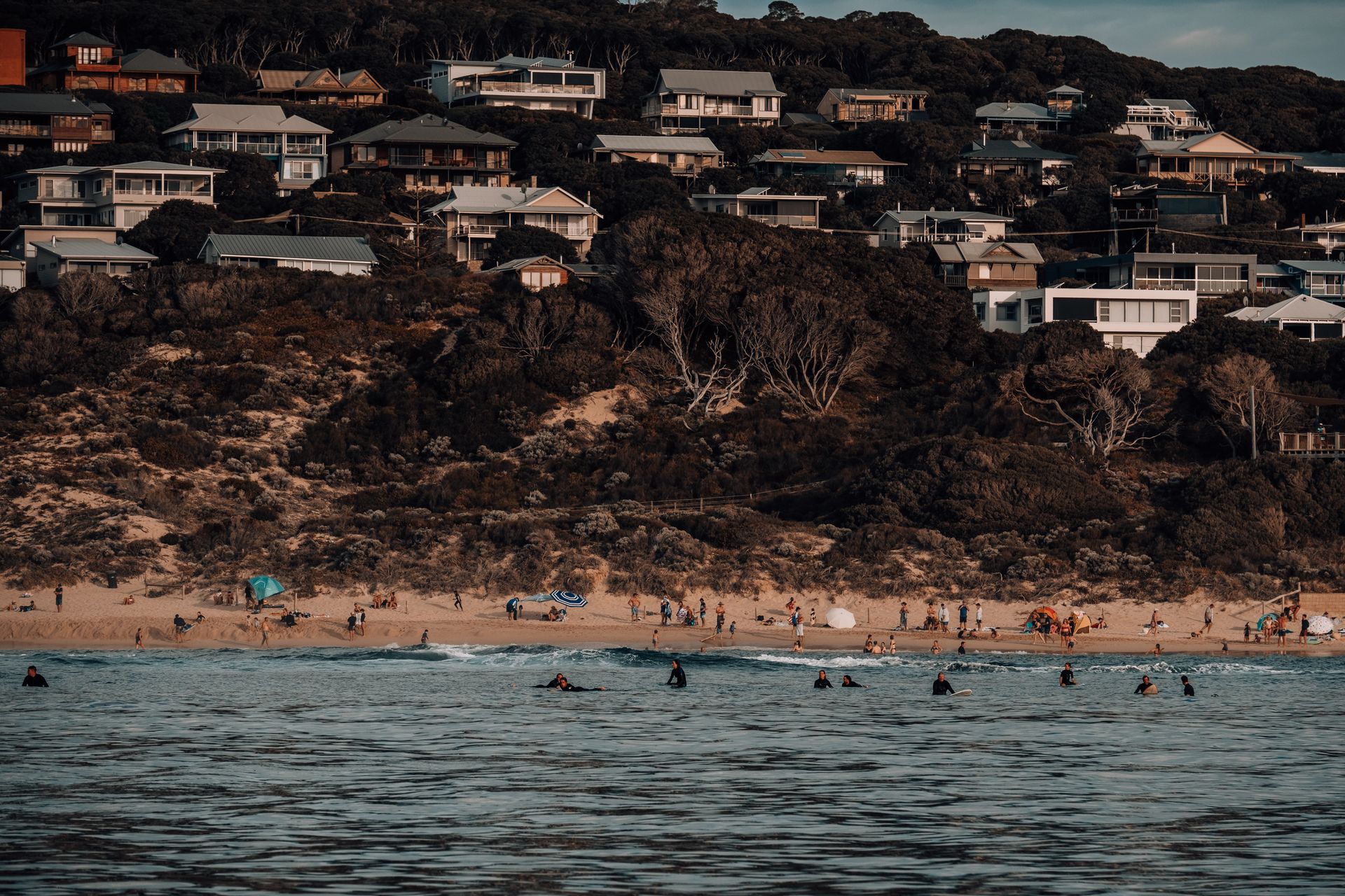 Idyllic beach scene with surfers and hillside vacation homes overlooking a sandy shore.