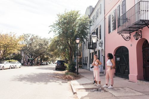 Free Two women stand on a vibrant urban street lined with pastel-colored buildings and trees. Stock Photo