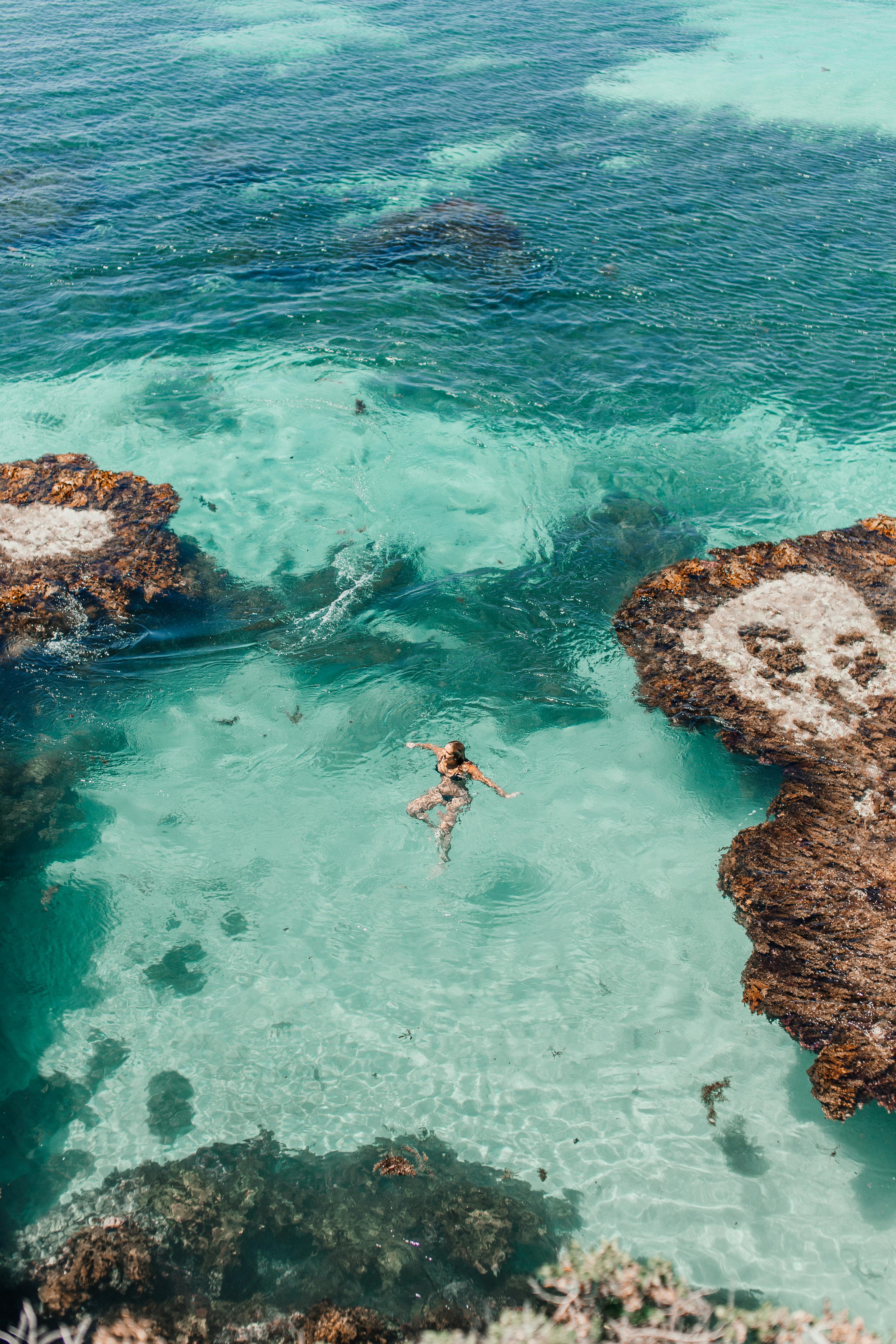 宁静的鸟瞰图，一名女子在清澈的碧绿海水中游泳，周围环绕着岩石