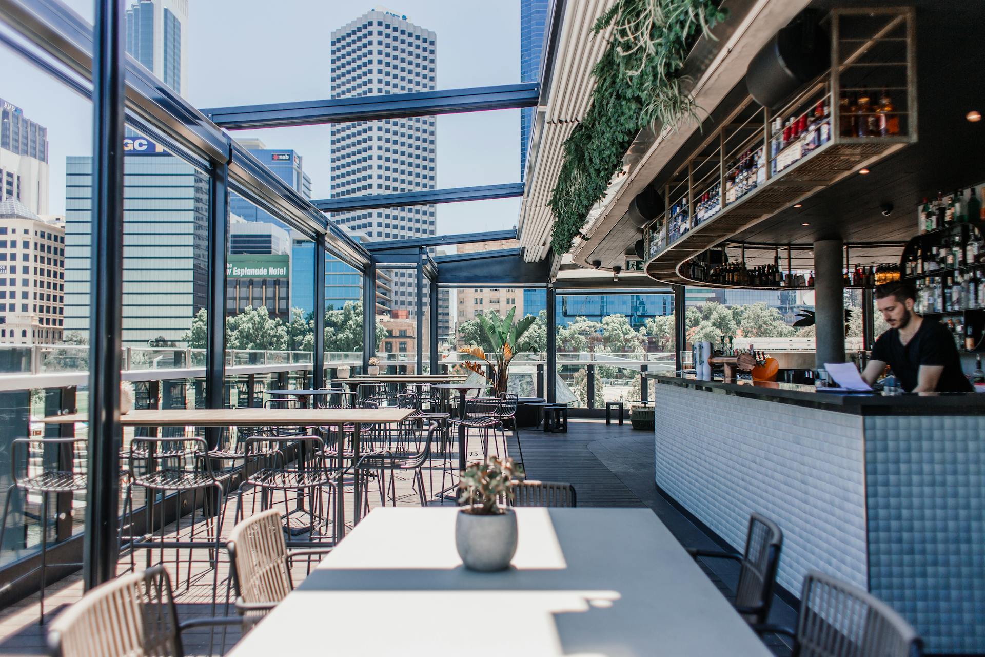 A stylish rooftop bar with city views featuring tables, chairs, and greenery.