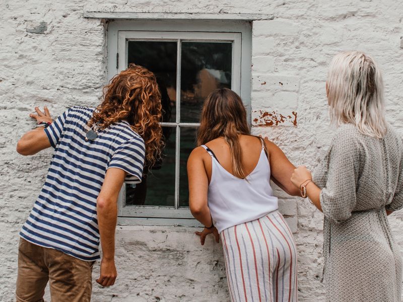 Three friends curiously peek through a vintage building window, exploring outdoors - historic homes
