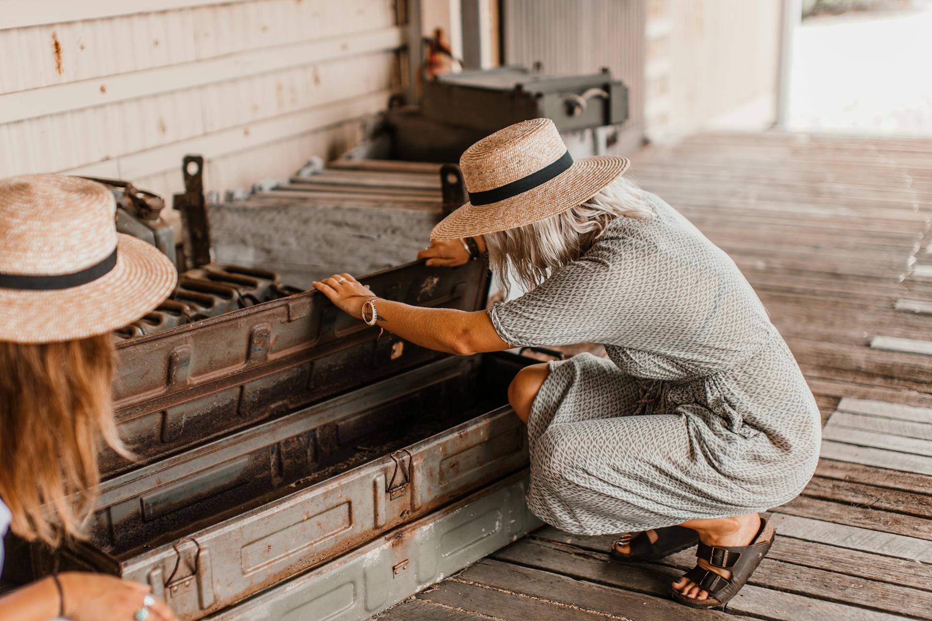 Two women in straw hats and dresses checking antique equipment outdoors.