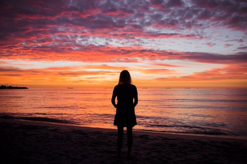 Free A woman stands on a beach, silhouetted by a vibrant pink and orange sunset over the ocean. Stock Photo