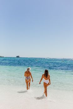 Two women in swimsuits walking along a sunny beach, enjoying the clear blue sea and sky.