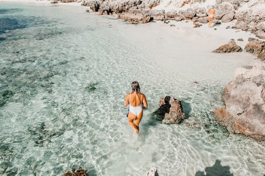 A woman walks on a rocky beach with clear waters and sunlight.