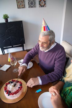 A senior man wearing a birthday hat cutting a cake indoors surrounded by party decor.