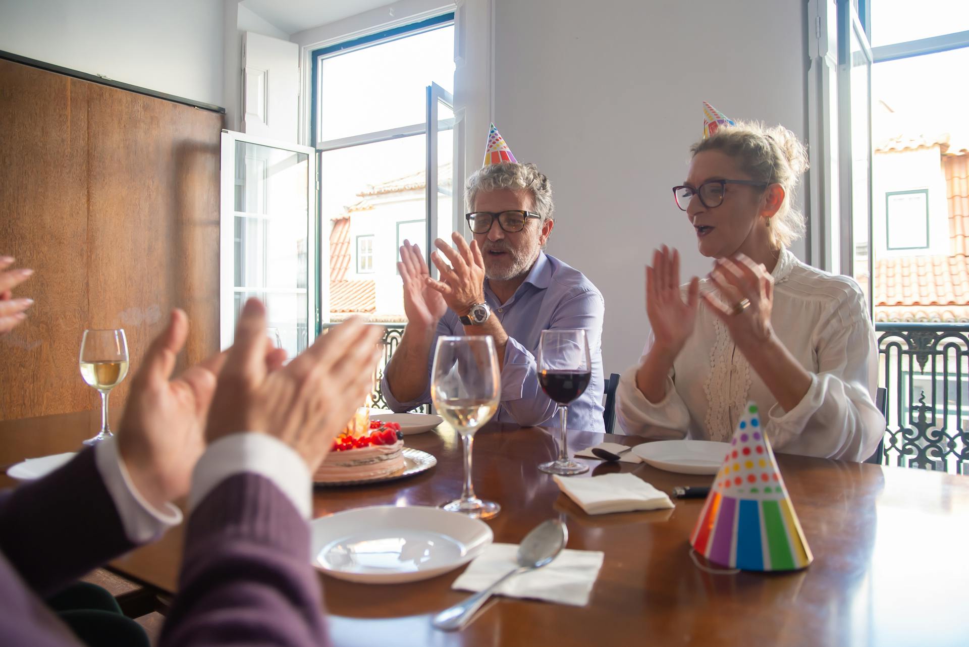 A senior couple celebrates a birthday indoors with friends, wearing party hats and enjoying cake.