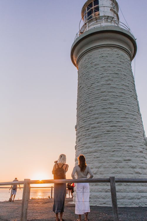 Free Two tourists observing sunset near lighthouse by the sea, capturing travel moments. Stock Photo