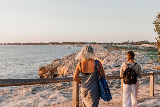 Two women walking along a scenic coastal path during sunset with a view of the rocky shoreline and sea.