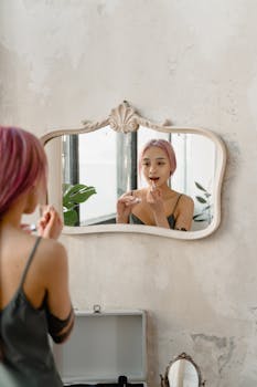 A young woman applying lip gloss in front of a decorative mirror inside a stylish room.