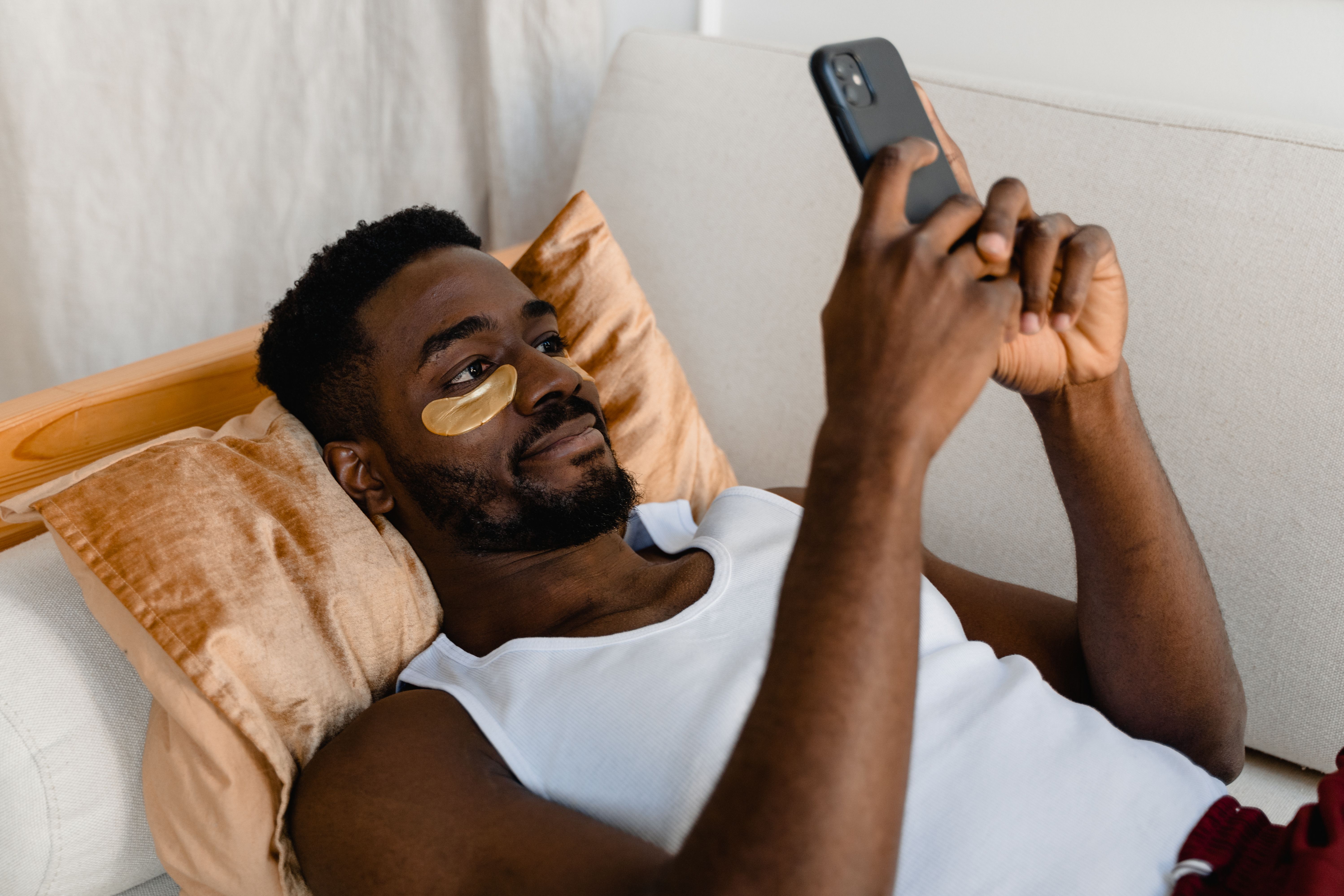 African American man lying down with eye patches, using a smartphone for relaxation and self-care.