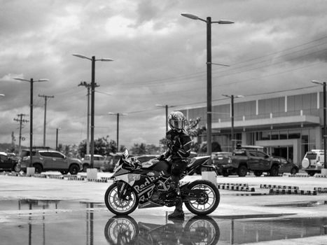 A motorcyclist in urban setting, reflecting on a wet asphalt in grayscale.