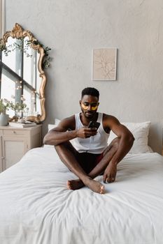 A young man with under-eye patches sitting on a bed, using a smartphone.