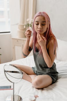 A woman with pink hair applies skincare while sitting on a bed, reflecting self-care.