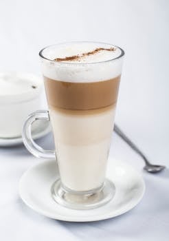 Close-up of a frothy latte macchiato served in a tall glass with a spoon on a white plate.