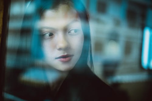 Free Through glass wall view of crop young ethnic female with makeup looking away in daytime Stock Photo