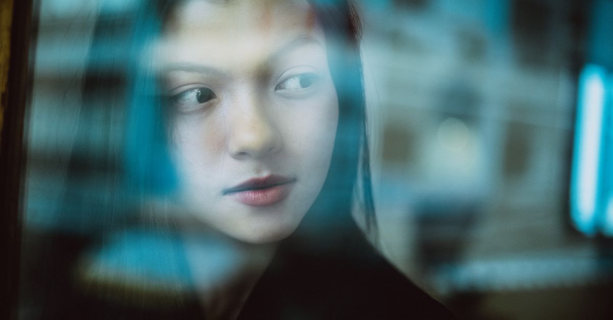 Through glass wall view of crop young ethnic female with makeup looking away in daytime