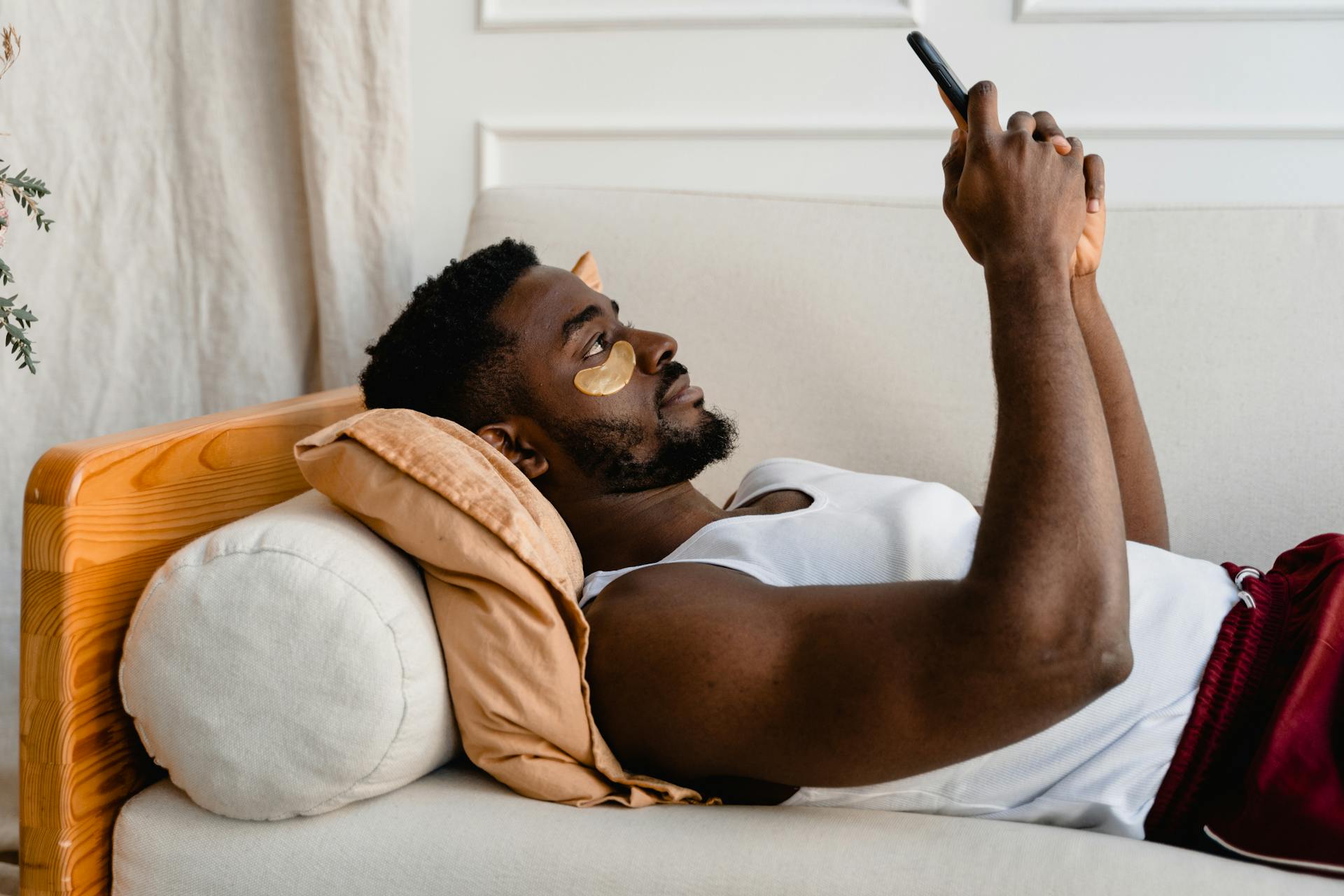 Black man lying on sofa, using smartphone, wearing under eye patches for self care and relaxation.