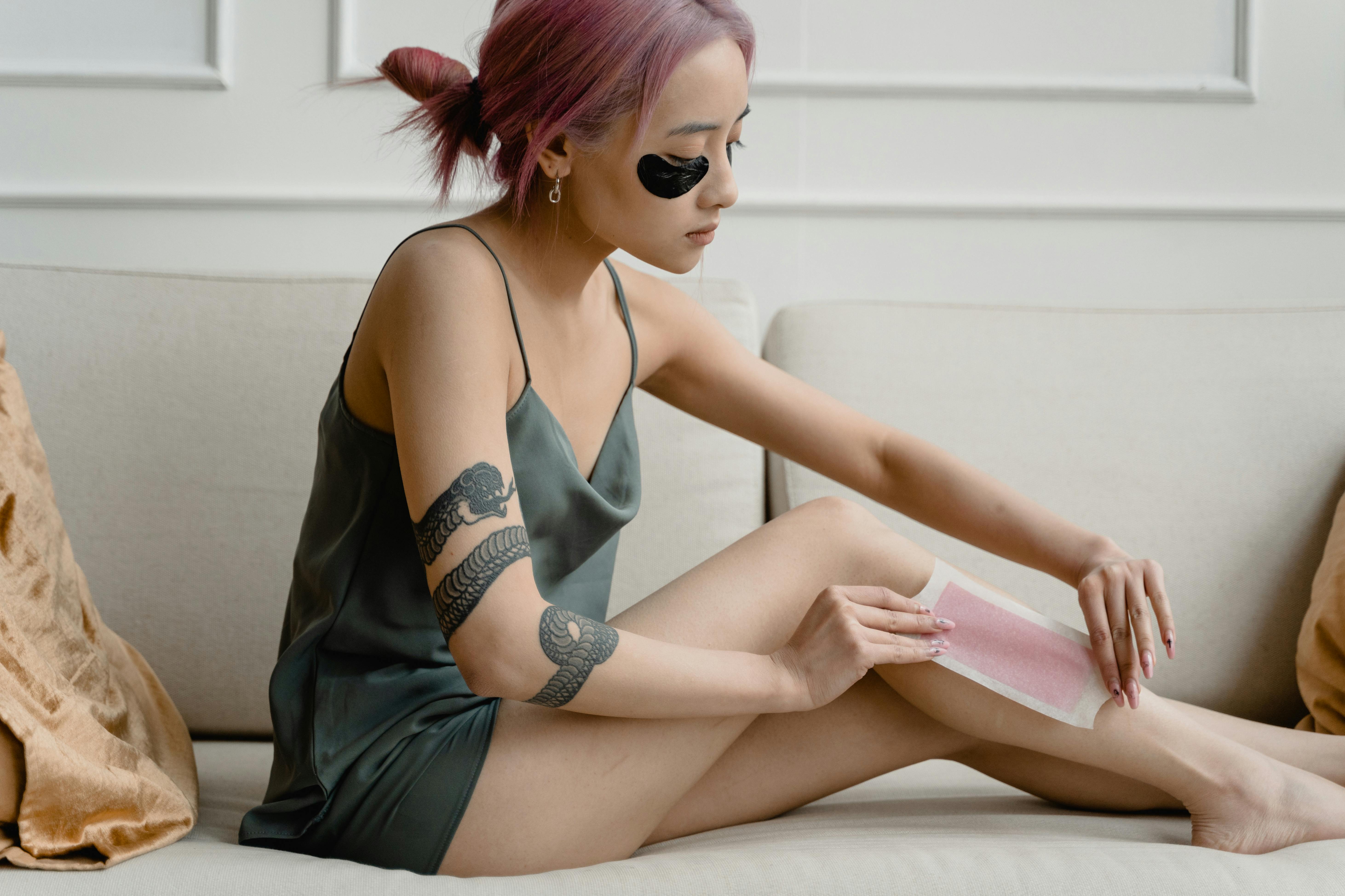A young woman with pink hair applies a waxing strip indoors, highlighting self-care and relaxation.