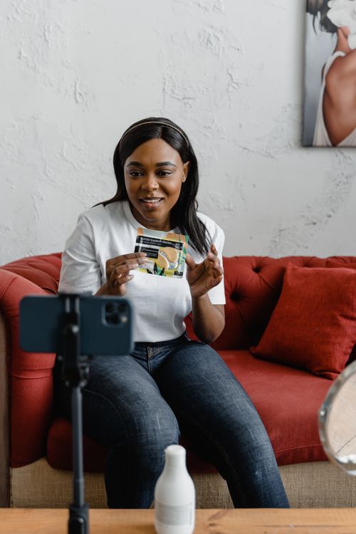 Free A woman sits on a red sofa showing a beauty product during a video call at home. Stock Photo