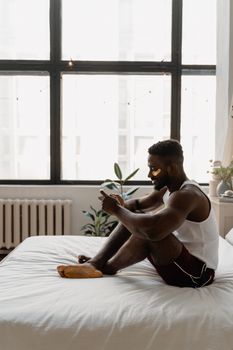 A man in a white tank top enjoys a relaxing moment with under-eye masks on a bed in a bright, modern bedroom.