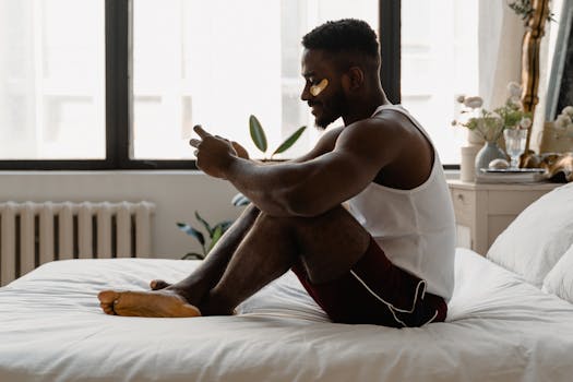 African American man sitting on bed, smiling with under eye patches, enjoying a skincare moment.