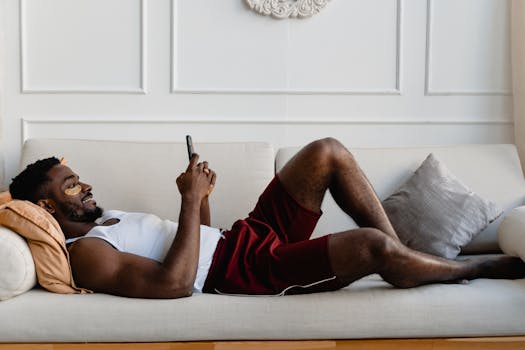 A man in casual wear enjoys skincare and smartphone time on a comfortable sofa at home.